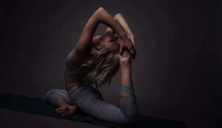 mid-30s woman in dark lit room arching back and holding feet in yoga pose battling the swerve